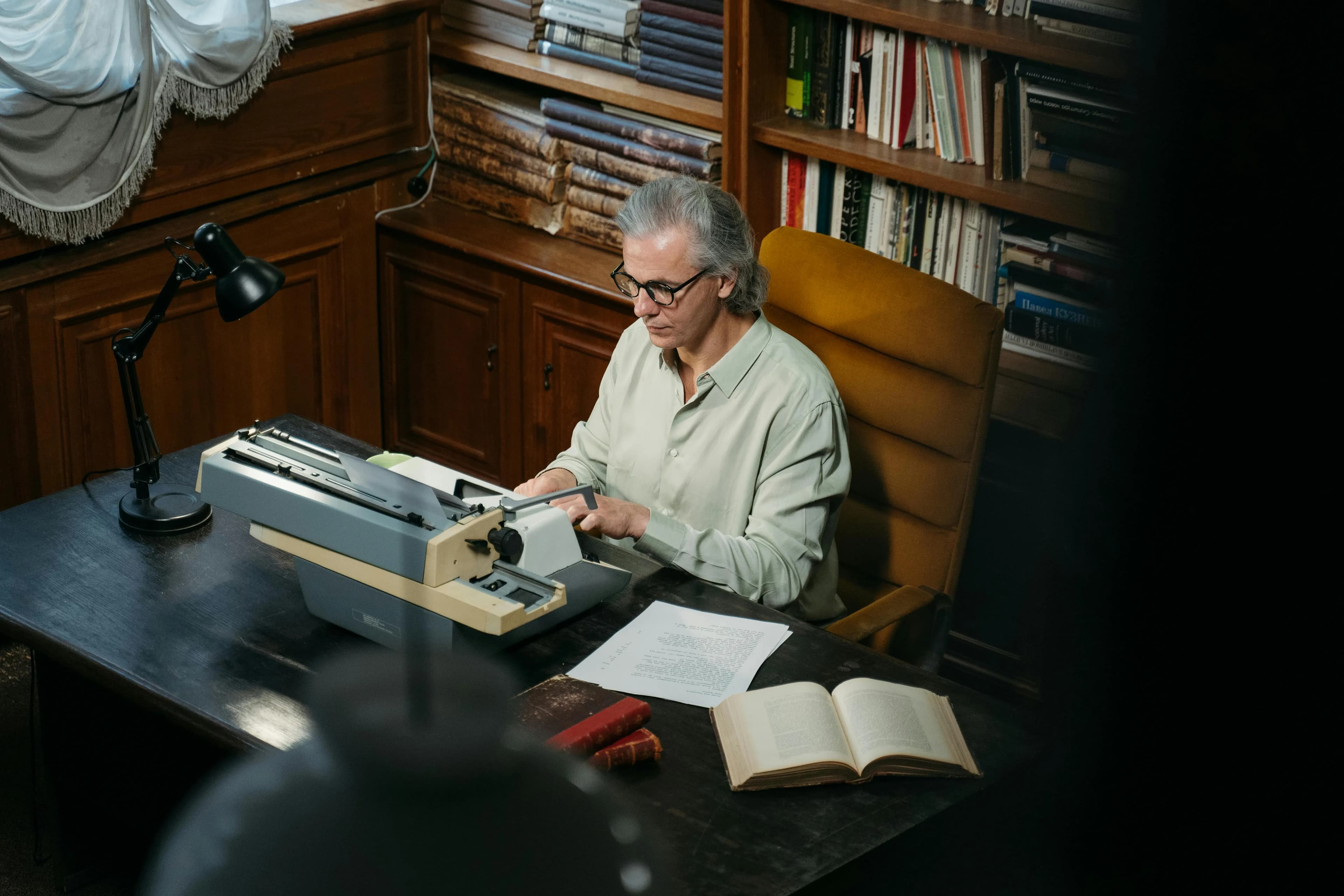 A man with glasses typing on a vintage typewriter at a wooden desk in a library filled with books.