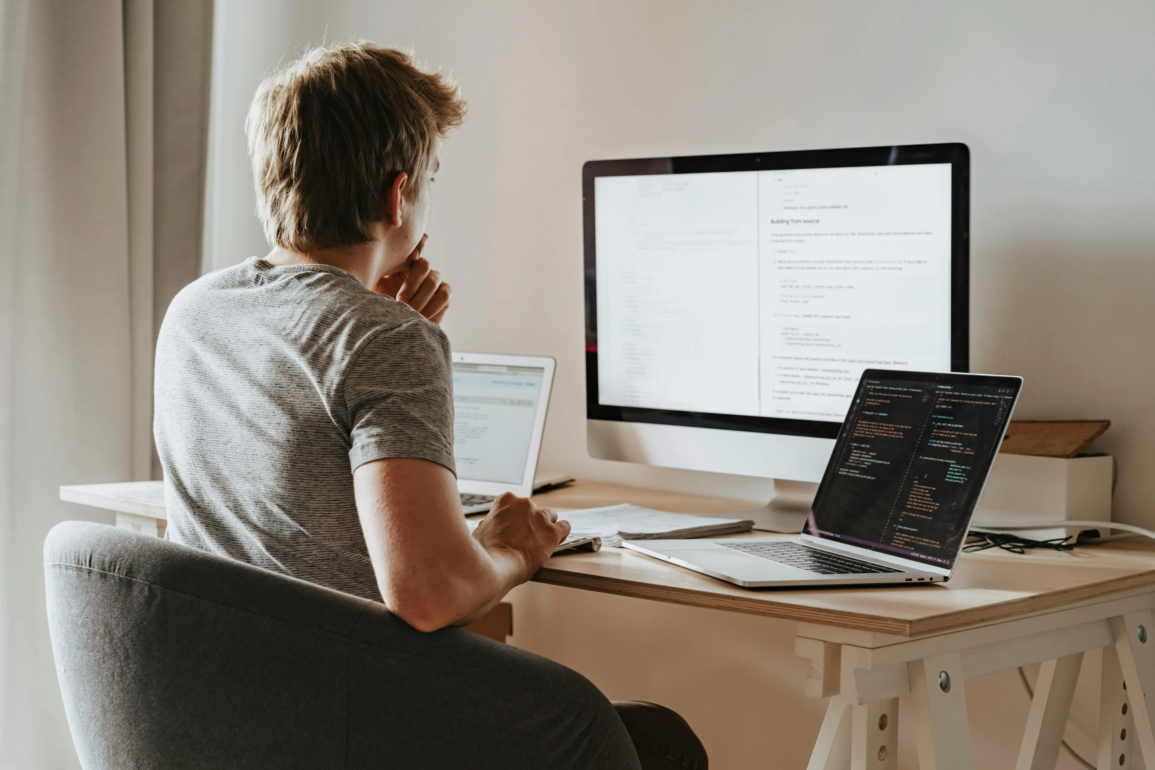 A developer seen from behind, hand to chin, reading code on a laptop and a large monitor