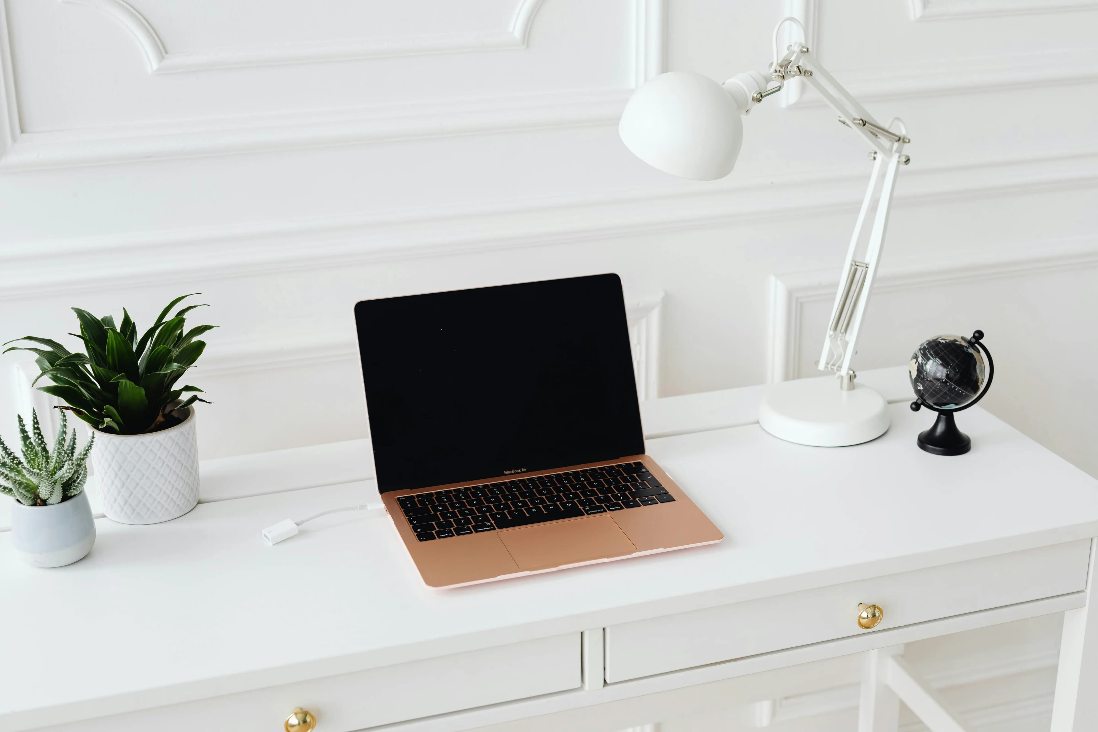 An open laptop on a white desk with a potted plant, an angled lamp and a small globe