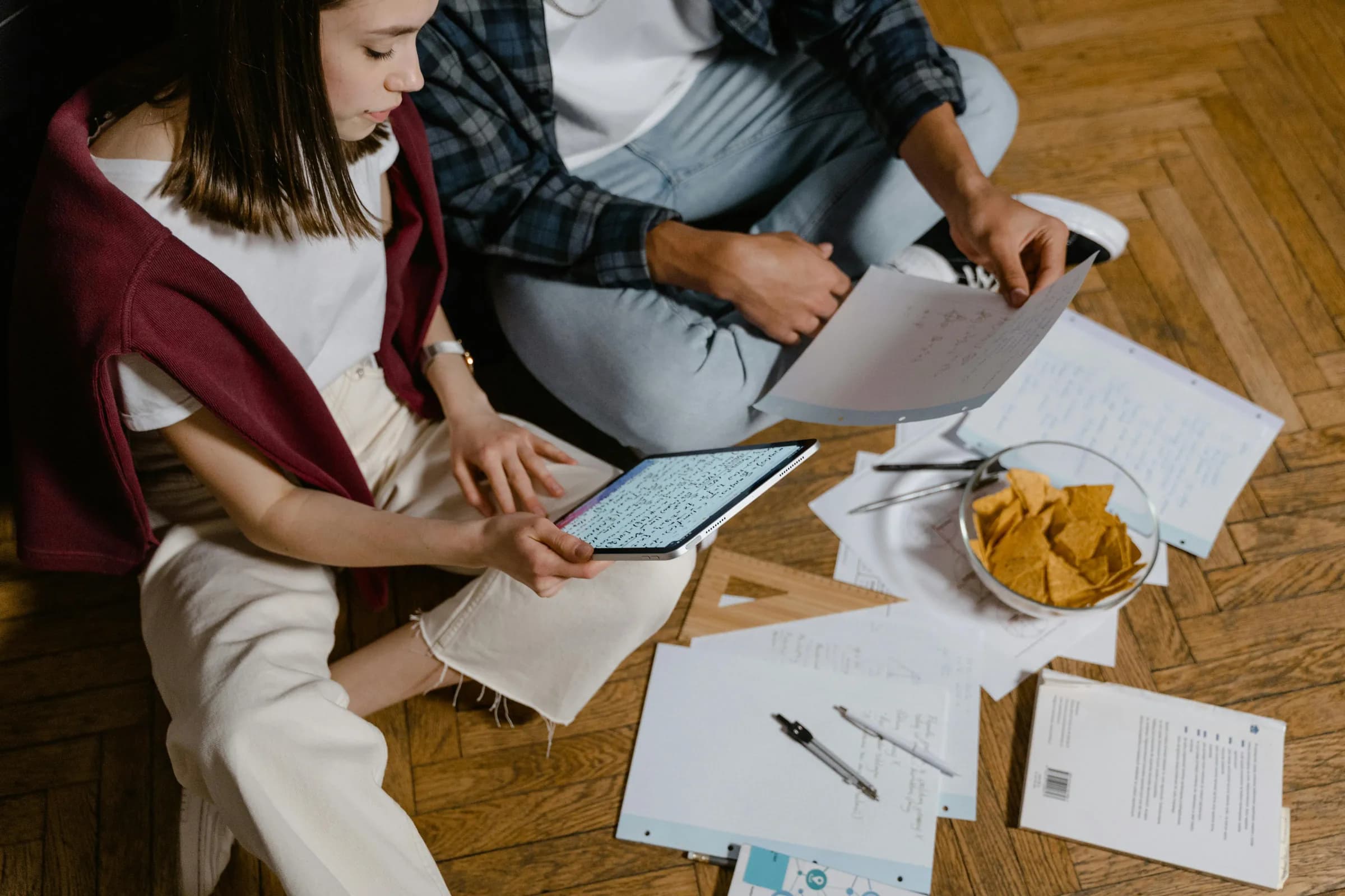 Two students sitting on a wooden floor studying with a tablet and paper documents
